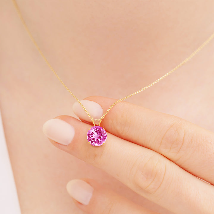 14K yellow gold lab-created pink sapphire pendant, bright candy pink, held between fingers for size reference.