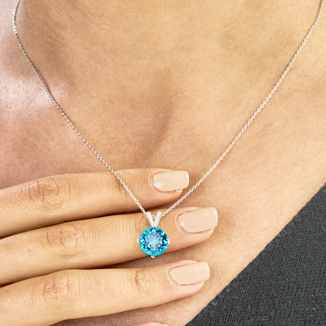 Hand holding an 8mm round Swiss blue topaz pendant necklace in sterling silver, with 4-prong setting and rolo chain.
